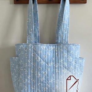 May include: A light blue quilted tote bag featuring a white floral pattern, two long shoulder straps, and two side pockets. The bag has vertical stitching throughout and includes a logo with a needle icon and the text Vamos Digital Sewing.