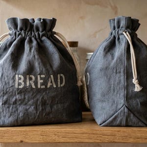May include: Two dark gray linen drawstring bags with a ruffled top edge and white cotton cords. The left bag features the word BREAD printed in a distressed white font. The bags have a structured base and are made of a textured fabric.