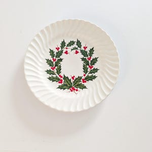 May include: White ceramic plate with a wavy rim design. The plate features a wreath of green holly leaves and red berries in the center. The plate is a festive holiday or Christmas themed item.