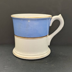 May include: A white ceramic mug with a blue band and gold trim. The mug has a handle and a rounded base.