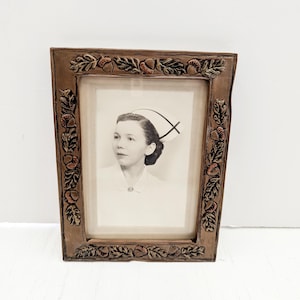 Pode incluir: Uma moldura de madeira vintage com um design de bolota e folha esculpido. A moldura contém uma fotografia em preto e branco de uma mulher vestindo um uniforme de enfermeira.