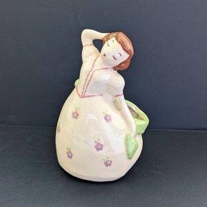 Puede incluir: Un macetero de cerámica con forma de mujer con cabello castaño, que lleva un vestido blanco con flores rosas y azules. El macetero tiene un asa en forma de hoja verde.