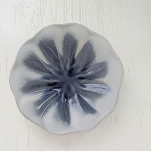 May include: A small, round, ceramic dish with a petal-like design. The dish is primarily gray with darker gray accents radiating from the center. The dish is sitting on a white, textured surface.
