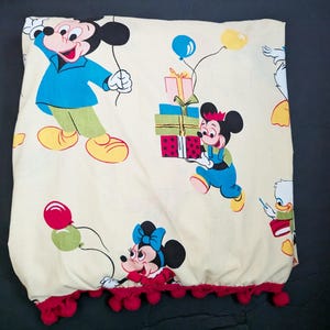 May include: A cream-colored fabric with a red pom-pom trim. The fabric features a repeating pattern of Mickey Mouse and Minnie Mouse characters, along with presents and balloons.