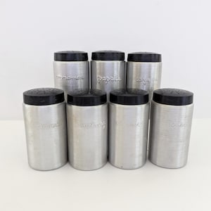 Puede incluir: Un conjunto de siete especieros vintage de aluminio con tapas de plástico negro. Cada especiero está etiquetado con el nombre de una especia en una fuente clásica. Los especieros están dispuestos sobre una superficie blanca.