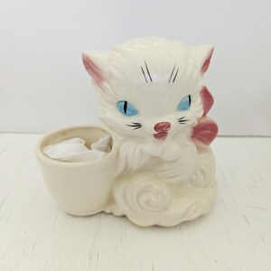 Puede incluir: Una figurita de gato de cerámica blanca con ojos azules y un lazo rosa. El gato sostiene una pequeña taza blanca en sus patas.