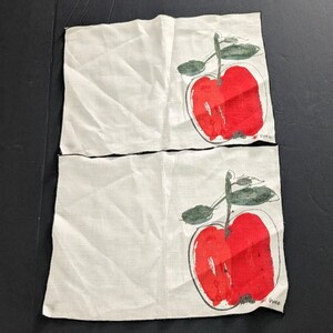 May include: Two white linen tea towels with a red apple design. The apples are stylized and have a green stem and leaves. The towels are hemmed with black thread.