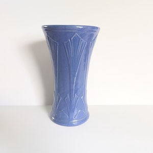 Puede incluir: Un jarrón de cerámica azul bígaro con forma acampanada. El jarrón presenta un diseño de hojas de estilo Art Déco repetido, tallado en la superficie. El jarrón mide aproximadamente 25 cm de altura.