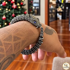 Puede incluir: Una pulsera de metal plateado con forma de dragón, que presenta una cabeza detallada con un ojo de piedra azul y un cuerpo texturizado similar a escamas que se envuelve alrededor de la muñeca. El diseño incluye una sección de cola segmentada y un acabado metálico.