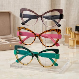 May include: Three pairs of cat-eye glasses displayed on a clear acrylic stand. The top pair has a brown tortoiseshell frame, the middle pair features a leopard print frame with pink temples, and the bottom pair has a green marbled frame.