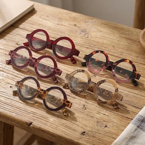 May include: Five pairs of round-framed glasses are arranged on a wooden surface. The collection includes one pair in solid dark red, two pairs with brown and black leopard print patterns, one pair in tortoiseshell, and one pair in light tan.