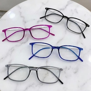 May include: Four pairs of rectangular spectacles with clear lenses and thin frames. The frames are available in four distinct colours: solid black, vibrant magenta, deep royal blue, and a translucent light grey, all featuring thin metal temples.