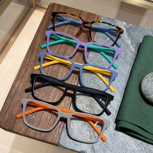 May include: A stack of five pairs of eyeglasses with rectangular frames. The top pair is tortoiseshell with blue temples, followed by purple frames with green temples, blue frames with yellow temples, black frames, and gray frames with orange temples.