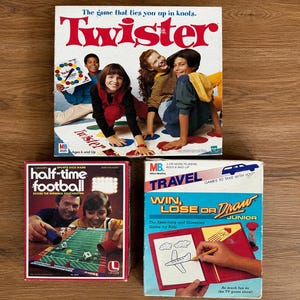 May include: Three board game boxes including Twister with a colorful mat, Half-Time Football featuring a green field graphic, and Travel Win, Lose or Draw Junior with a red drawing board illustration. Each box displays game titles and imagery.
