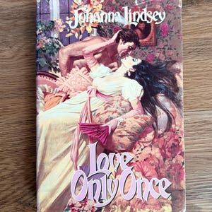May include: A paperback book titled Love Only Once by Johanna Lindsey. The cover features a romantic illustration of two figures in historical attire, with a woman in a cream dress holding a pink fan and a man leaning over her amidst floral decor.