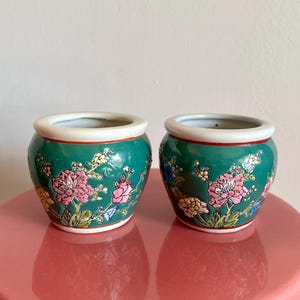 May include: Two small, round ceramic pots with a glossy teal finish. Each pot features a white rim, a red band, and a floral design with pink, yellow, and blue flowers. The pots are displayed on a pink surface.