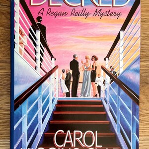 May include: A book cover with a pink and orange sunset background. The title "DECKED" is in large white letters. The subtitle "A Regan Reilly Mystery" is in smaller white letters. The author's name "CAROL HIGGINS CLARK" is in large white letters at the bottom of the cover. The cover features a group of people standing on a ship's deck with a staircase leading down.