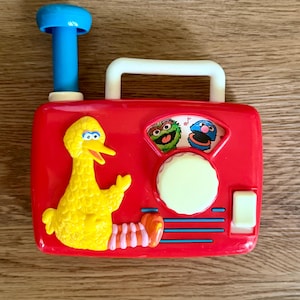 May include: A red plastic toy radio featuring a yellow Big Bird figure on the left side. The radio has a white handle, a blue antenna, a large round white dial, and a small white rectangular button. A graphic of Oscar the Grouch and Cookie Monster is visible above the dial.