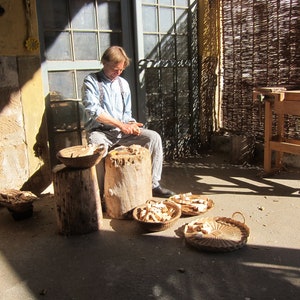 May include: A man is carving wood with a knife, sitting on a tree stump. There are several baskets filled with wood shavings around him.