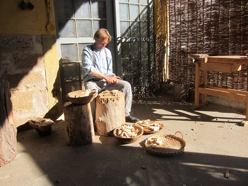 May include: A man is carving wood with a knife, sitting on a tree stump. There are several baskets filled with wood shavings around him.