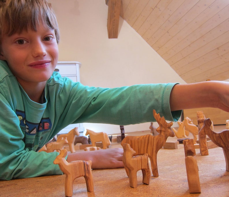 May include: A young boy plays with wooden animal figurines, including a deer, a horse, and a cow. The figurines are hand-carved and have a natural wood finish.