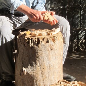 May include: A person carving wooden shapes with a knife. The shapes are scattered on a tree stump. There is a basket of additional wooden shapes in the foreground.