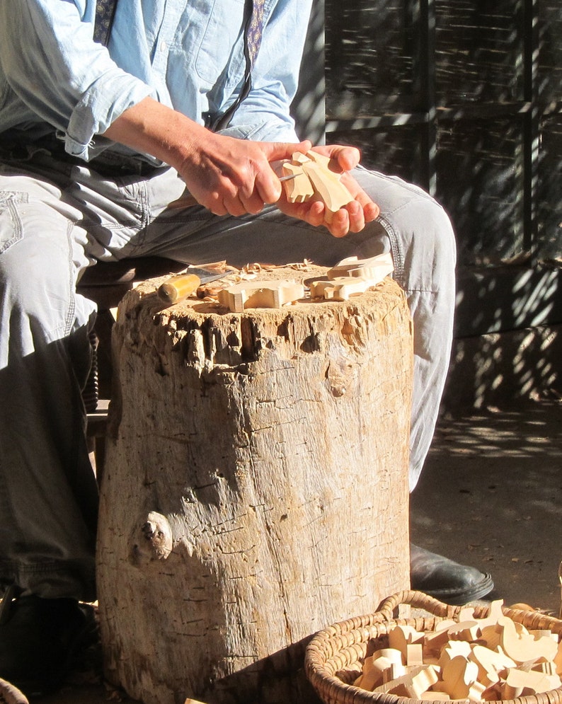 May include: A person carving wooden shapes with a knife. The shapes are scattered on a tree stump. There is a basket of additional wooden shapes in the foreground.