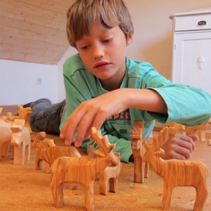 May include: A young person is playing with wooden animal figurines. The figurines are carved from wood and have a natural finish. The figurines include a deer, a giraffe, and a horse. The figurines are arranged on a wooden floor.