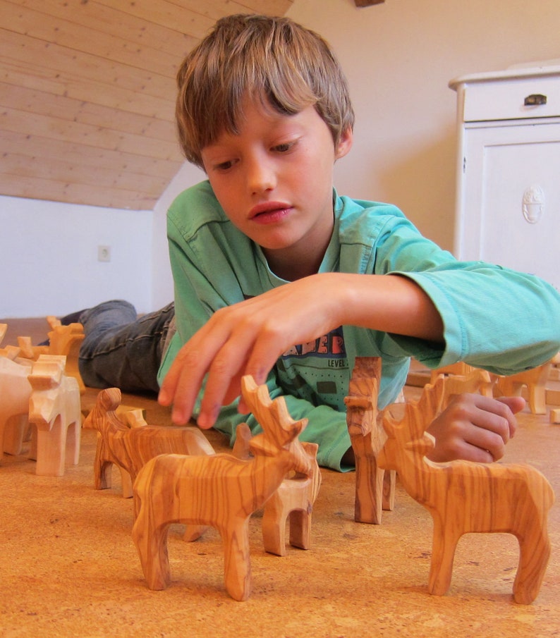 May include: A young person is playing with wooden animal figurines. The figurines are carved from wood and have a natural finish. The figurines include a deer, a giraffe, and a horse. The figurines are arranged on a wooden floor.