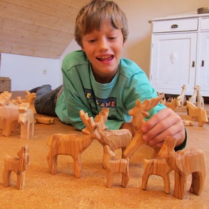 May include: A young person plays with wooden animal figurines, including a deer, moose, and a smaller fawn. The figurines are carved from wood with a natural wood grain finish. The figurines are arranged on a light brown surface.