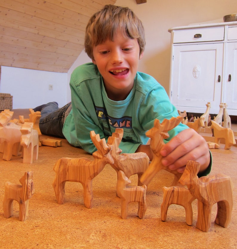 May include: A young person plays with wooden animal figurines, including a deer, moose, and a smaller fawn. The figurines are carved from wood with a natural wood grain finish. The figurines are arranged on a light brown surface.