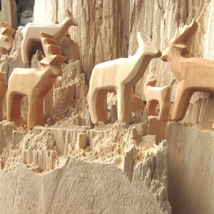May include: A group of wooden deer figurines carved from a single piece of wood. The deer are arranged in a cluster, with some standing upright and others lying down. The wood is a light brown color and has a rough, textured surface.
