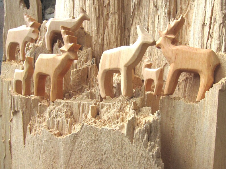 May include: A group of wooden deer figurines carved from a single piece of wood. The deer are arranged in a cluster, with some standing upright and others lying down. The wood is a light brown color and has a rough, textured surface.
