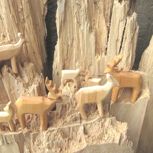 May include: A group of wooden deer figurines, carved in a simple style, are arranged on a piece of rough-hewn wood. The deer are all facing different directions, and some have antlers. The wood is a light brown color, and the deer are a slightly darker brown.