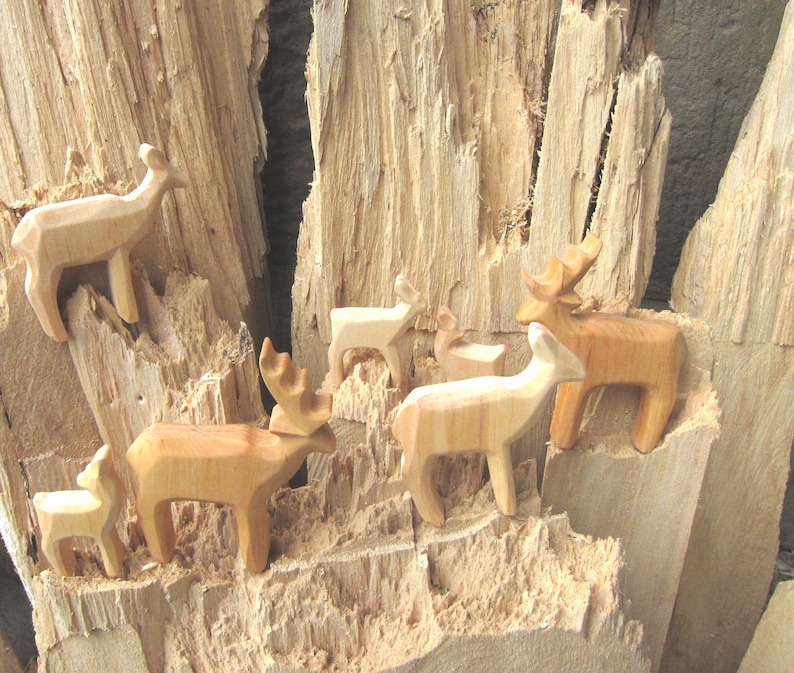 May include: A group of wooden deer figurines, carved in a simple style, are arranged on a piece of rough-hewn wood. The deer are all facing different directions, and some have antlers. The wood is a light brown color, and the deer are a slightly darker brown.