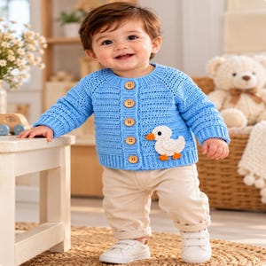 May include: A light blue long-sleeved knit cardigan featuring a row of five brown wooden buttons down the front and a white crocheted duck appliqué with an orange beak and feet on the lower right side, paired with beige trousers and white trainers.