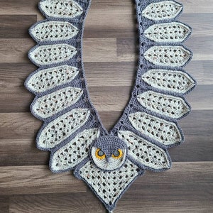 May include: A crocheted owl scarf in a grey and white colour scheme. The scarf features a large owl face in the centre with yellow eyes and a grey beak. The scarf is finished with a white tassel.