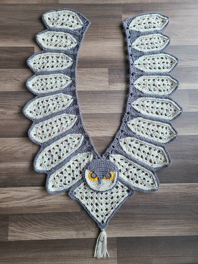 May include: A crocheted owl scarf in a grey and white colour scheme. The scarf features a large owl face in the centre with yellow eyes and a grey beak. The scarf is finished with a white tassel.