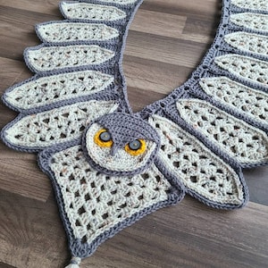 Könnte beinhalten: Gestrickte Eulen-Schal aus grauem und weißem Garn, mit einem detaillierten Eulengesicht mit gelben Augen. Der Schal hat eine lange Franse am unteren Ende.