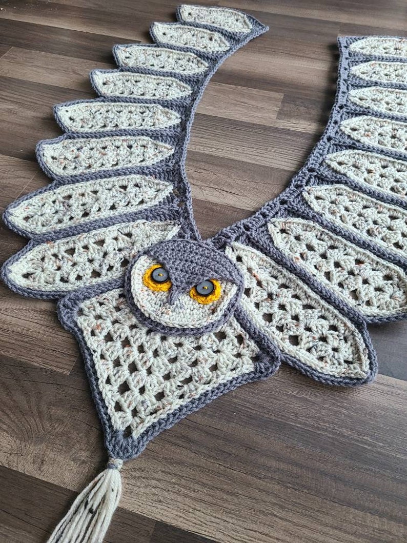May include: Crocheted owl scarf in grey and white yarn, featuring a detailed owl face with yellow eyes. The scarf has a long fringe at the bottom.