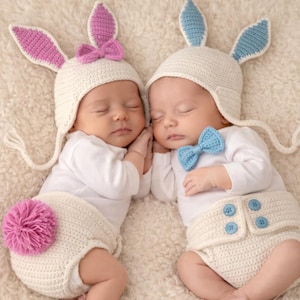May include: Two sleeping babies wearing white bunny hats with pink and blue accents, white onesies, and matching diaper covers. One has a pink bow and pom-pom, the other a blue bow.