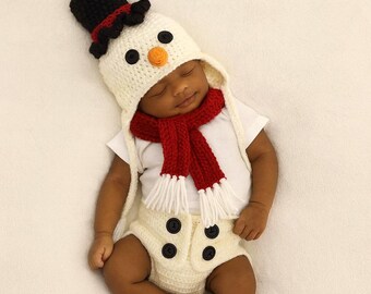 May include: A newborn baby dressed in a snowman costume. The costume includes a white hat with a black top and red trim, a red scarf with white fringe, and cream-colored diaper cover with black buttons. The baby is lying on a white surface.
