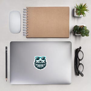 May include: A sticker featuring a cartoon illustration of a bearded man wearing blue sunglasses. The man has a black beard and moustache. The sticker is on a silver laptop.