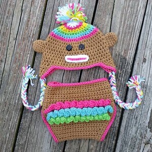 Può includere: Un set di cappello e pannolino all'uncinetto per un bambino scimmia. Il cappello ha una striscia arcobaleno e un pon pon bianco in cima. Il pannolino ha un bordo rosa, blu e verde.