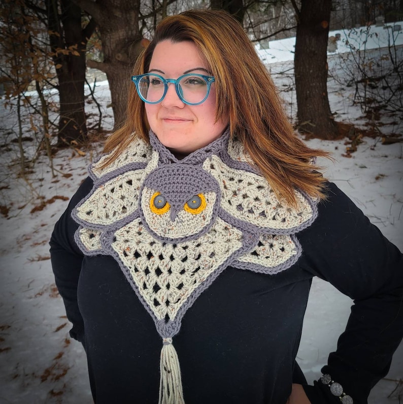 May include: A crocheted owl cowl with a grey owl face and yellow eyes. The cowl is made with a cream and grey yarn and has a long tassel.