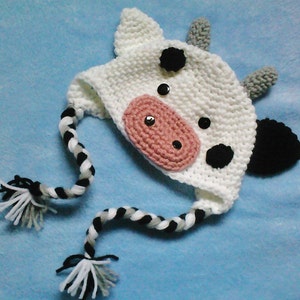 May include: White crocheted cow hat with black and white braided ties. The hat has black and white spots and a pink nose.