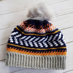 Könnte beinhalten: Eine graue, blaue, orange und weiße Häkelmütze mit einem weißen Kunstfellbommel oben.