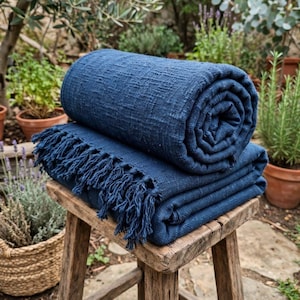 May include: A deep navy blue woven textile throw blanket with a textured surface and fringed edges. The blanket is folded and rolled, resting on a rustic wooden stool. The fabric features a visible cross-hatch weave pattern throughout the material.