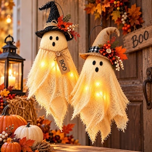 May include: Two hanging ghost decorations made of beige textured fabric, illuminated by internal lights. One wears a black hat, the other a burlap hat, both adorned with autumn leaves and berries. A small tag on the first ghost reads BOO.