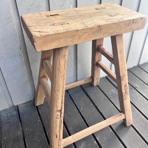 May include: A rectangular wooden stool with a light brown, weathered finish. The stool features a thick, solid top with visible wood grain and natural imperfections, supported by four sturdy, slightly angled legs connected by horizontal support bars.